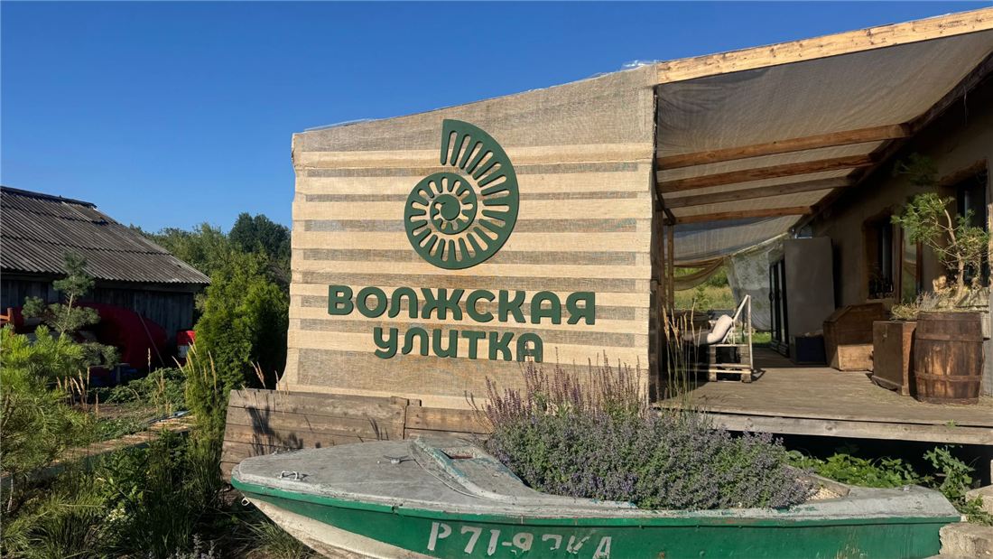 Вывеска «Волжская улитка» с ракушкой на стене из мешковины, рядом лодка с цветами